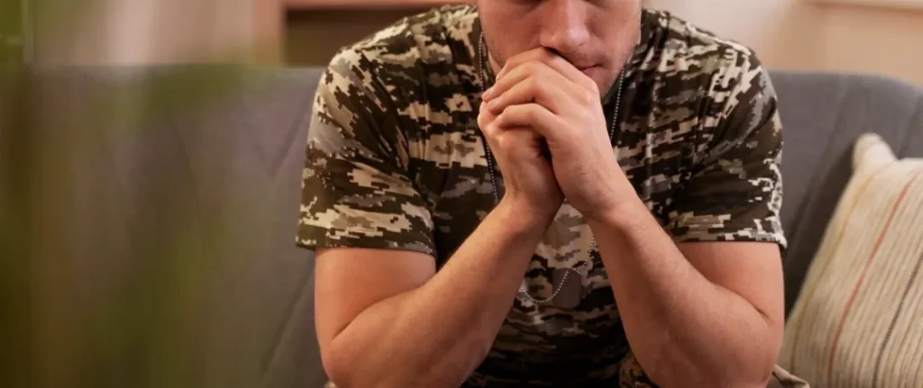 A man dressed in military camouflage clothes sits on a sofa with his hands on his chin, looking to the side thoughtfully. He is a former Marine who lived at Camp Lejeune and was exposed to the water contamination. He is part of the mass tort lawsuit that seeks accountability and compensation from the U.S. government.